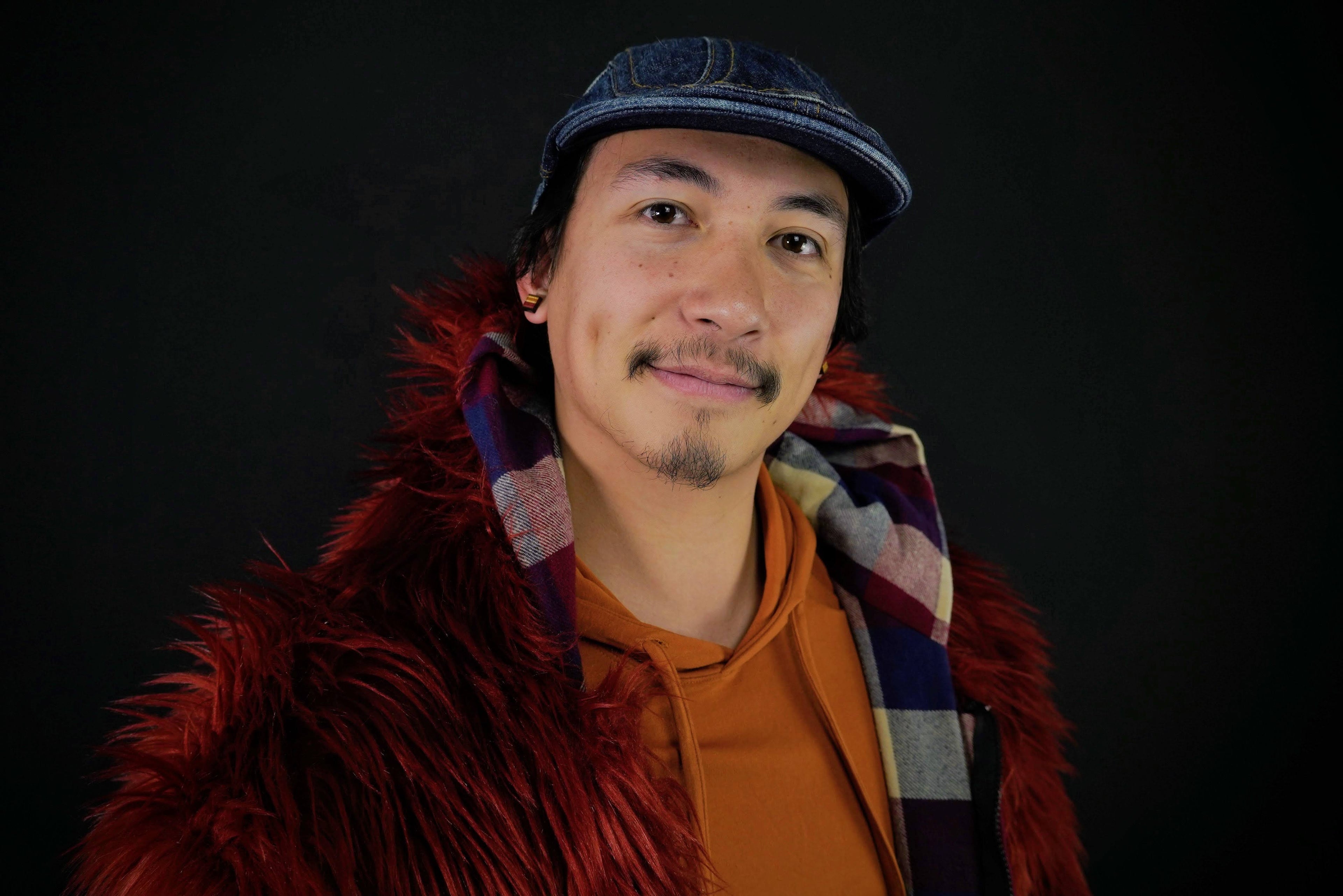 Man wearing a red fur coat, orange sweater, and plaid scarf against a black background